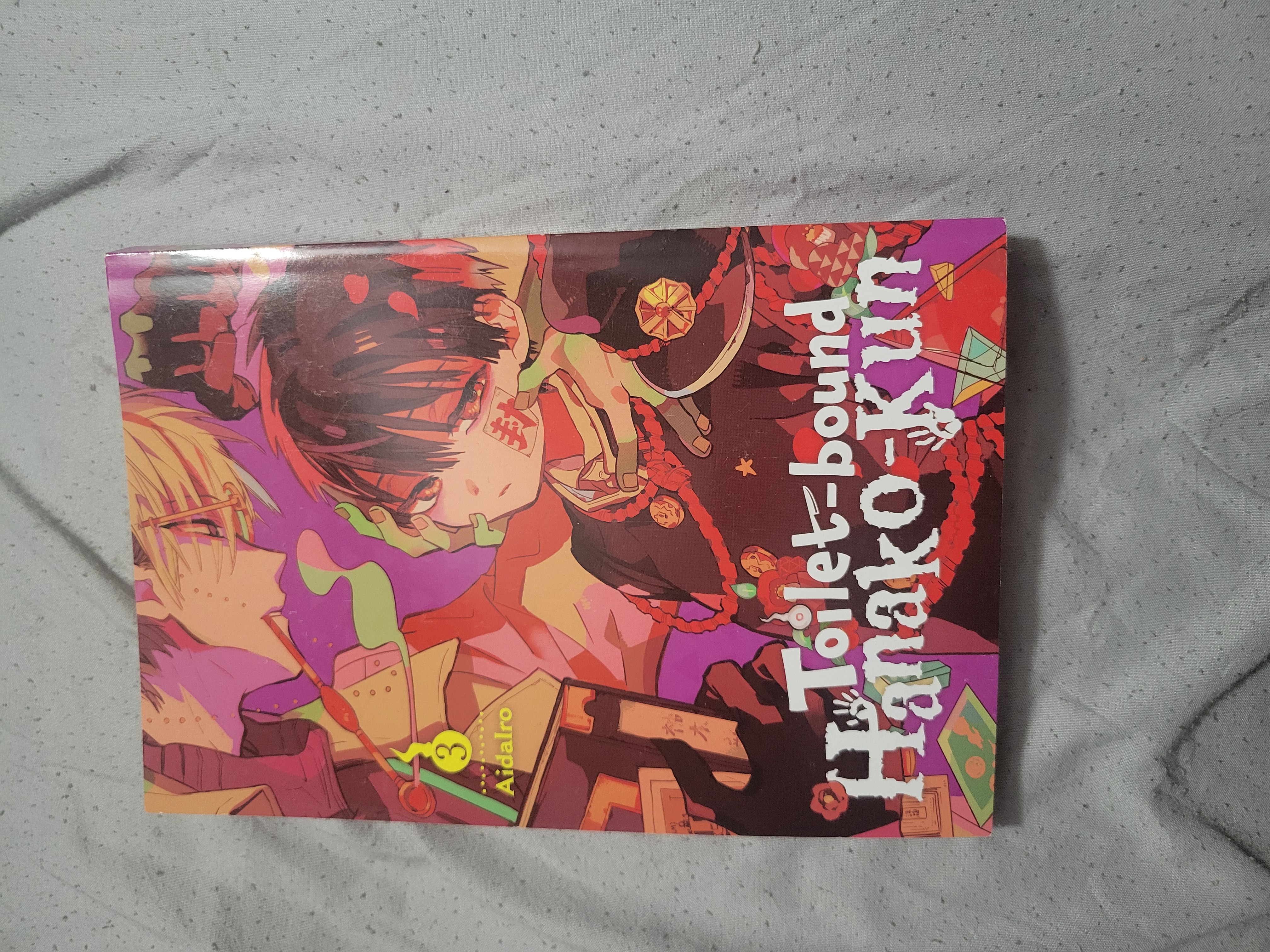 Front cover of Back of Toilet-bound Hanako-kun volume three on a gray bedsheet.